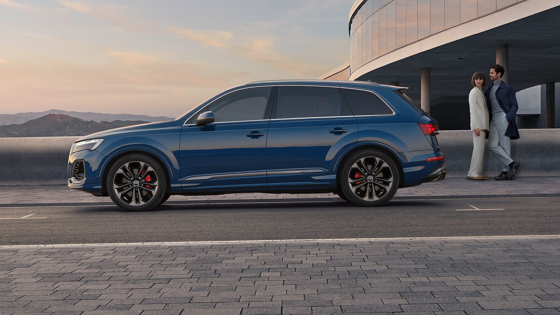  Seitenansicht eines blauen Audi Q7, eine Frau und ein Mann stehen hinter dem Fahrzeug.