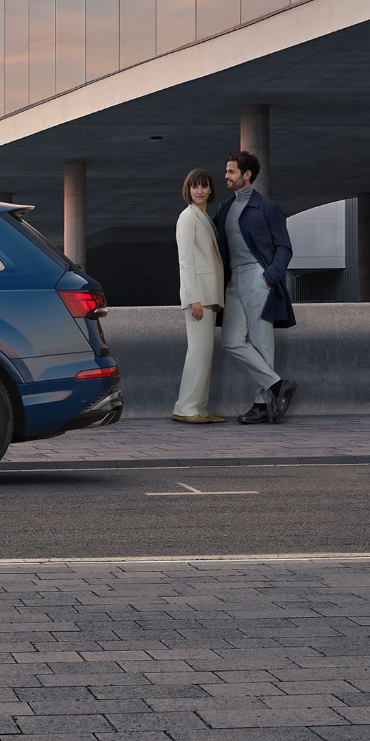  Seitenansicht eines blauen Audi Q7, eine Frau und ein Mann stehen hinter dem Fahrzeug.