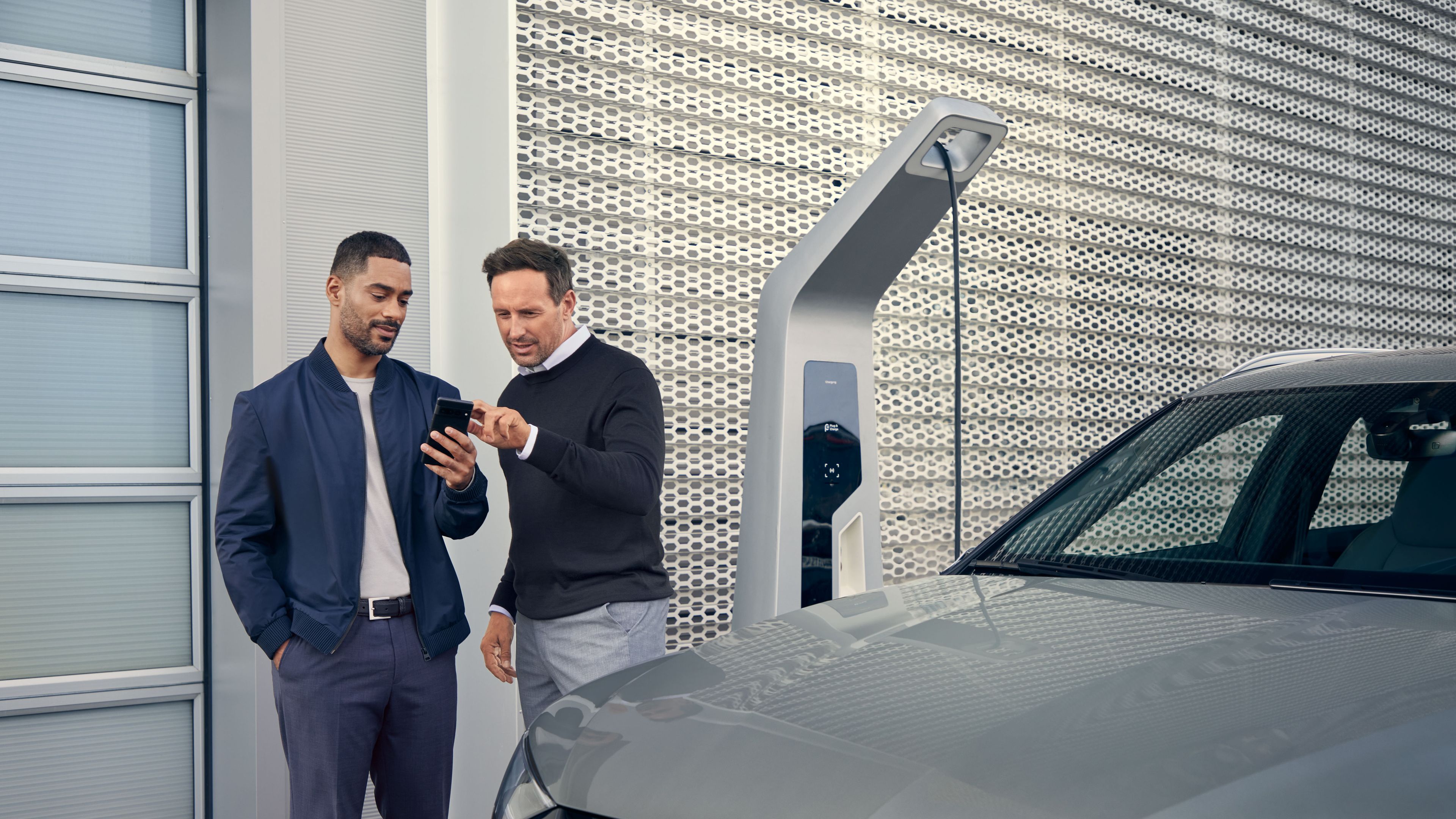 Ein Audi Service Mitarbeiter und ein Kunde blicken auf ein Smartphone, im Vordergrund steht ein Audi Elektroauto
