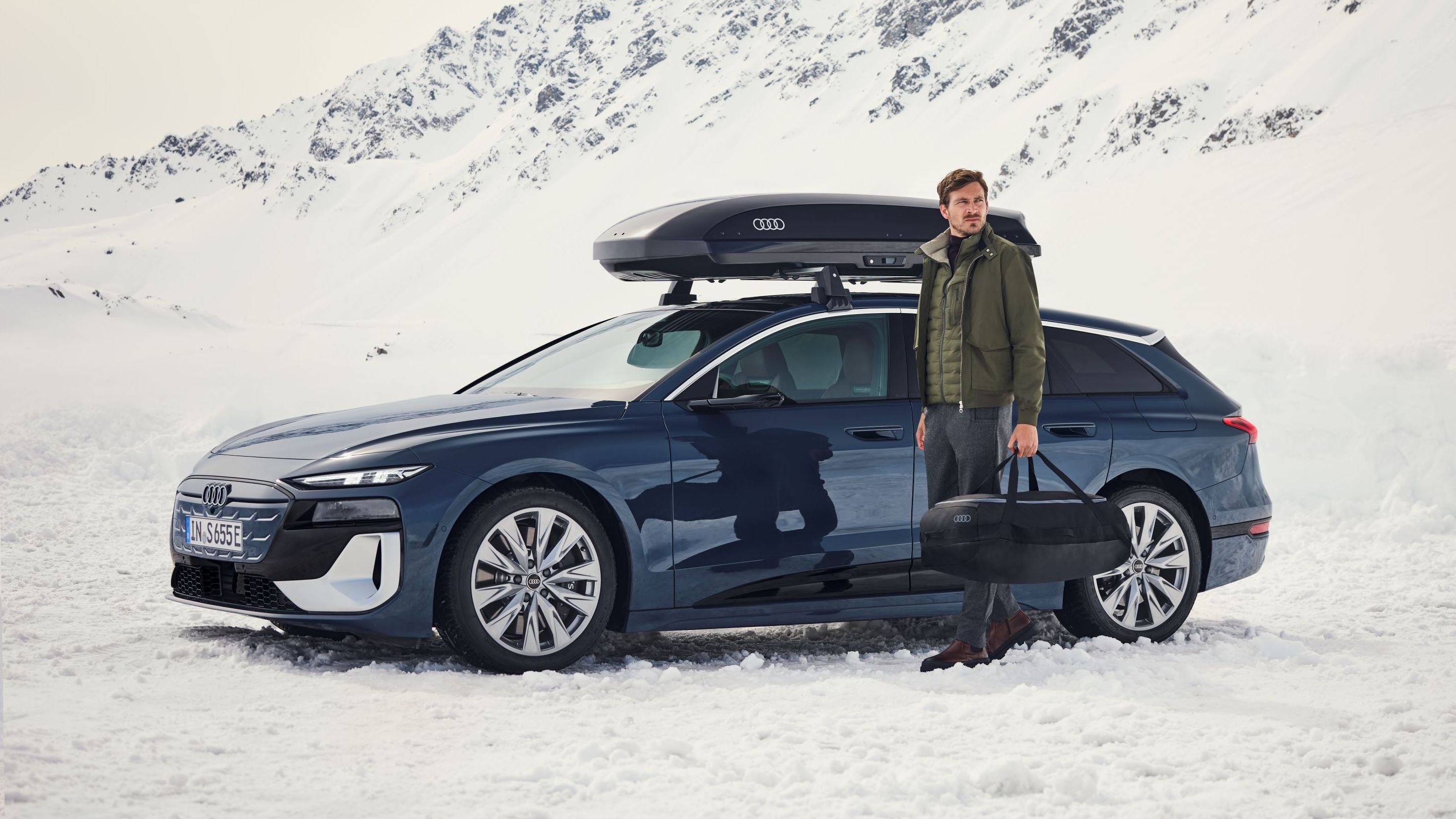 Ein Audi S6 mit Dachbox parkt in einer winterlichen Berglandschaft, ein Mann mit Tasche steht davor