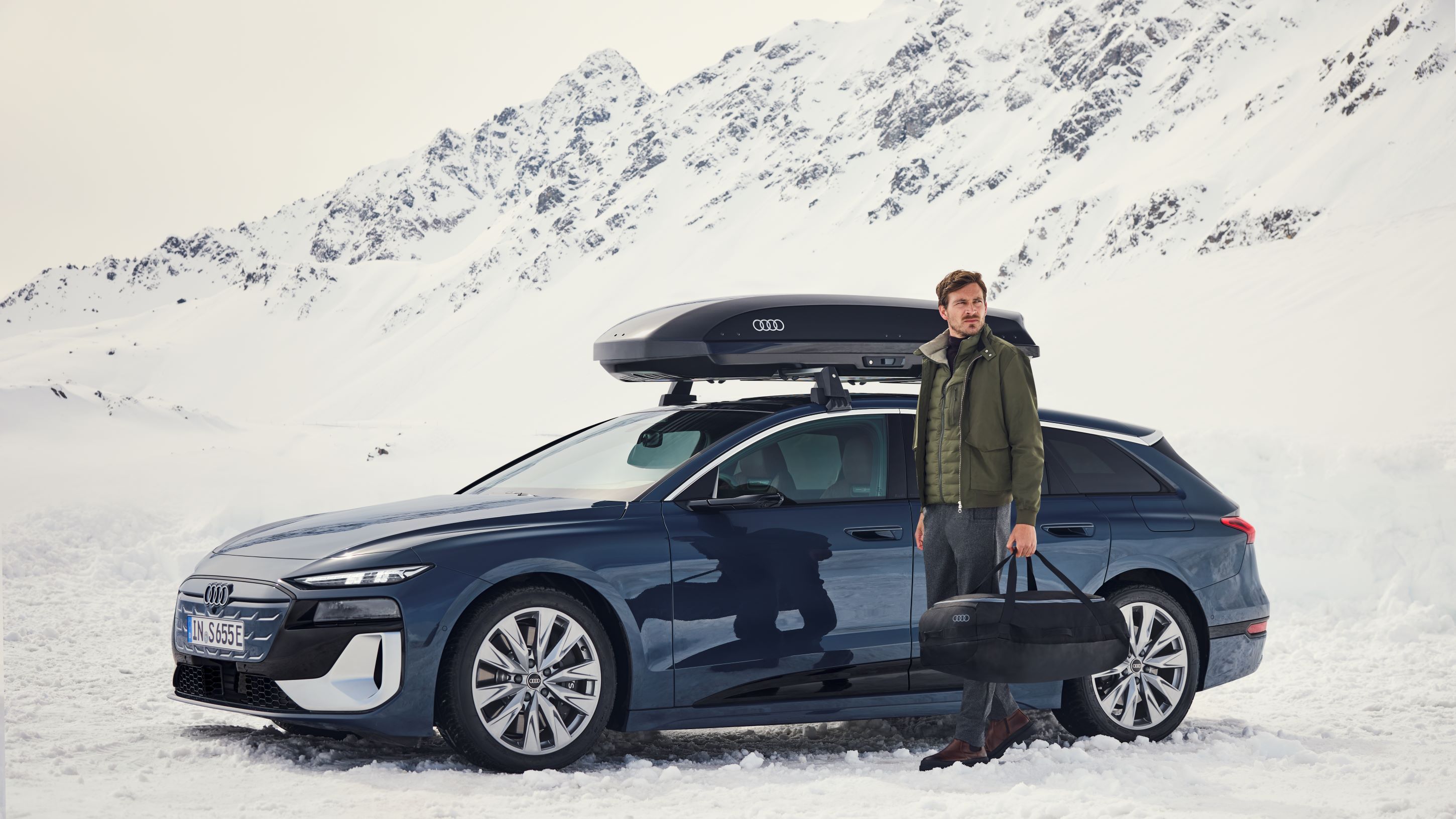 Ein Audi S6 mit Dachbox parkt in einer winterlichen Berglandschaft, ein Mann mit Tasche steht davor